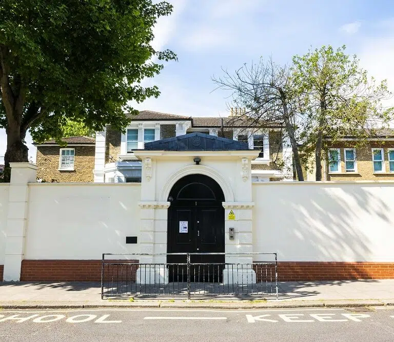 White perimeter wall with an arched entrance and black door leading into a school, with trees and a blue sky behind.