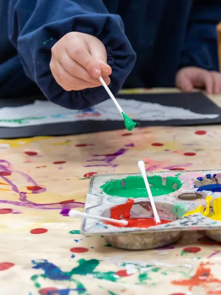 Child’s hand holds a brush with green paint above a mixed-color paint palette on a splattered tablecloth menagerie of colors.