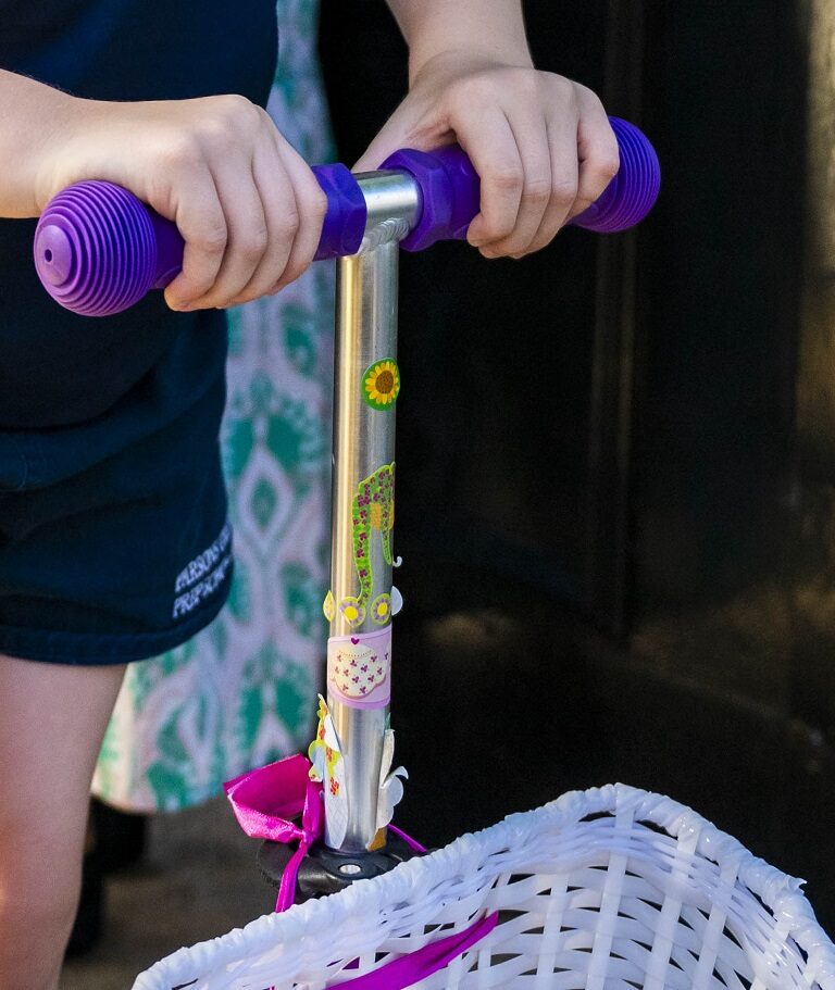 Child gripping purple scooter handlebars; silver stem decorated with stickers and a pink ribbon on a white wicker basket in front.