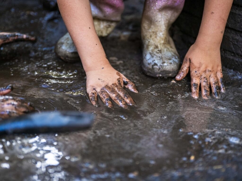 Child's hands, stained with mud, press into a wet, muddy ground outdoors with muddy boots nearby.