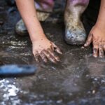 Child's hands, stained with mud, press into a wet, muddy ground outdoors with muddy boots nearby.