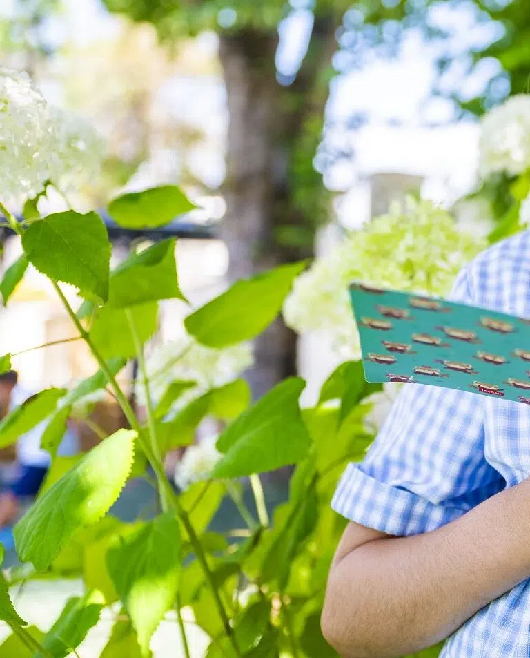 Person in a blue checkered shirt holds a teal brochure while standing among green plants outdoors.
