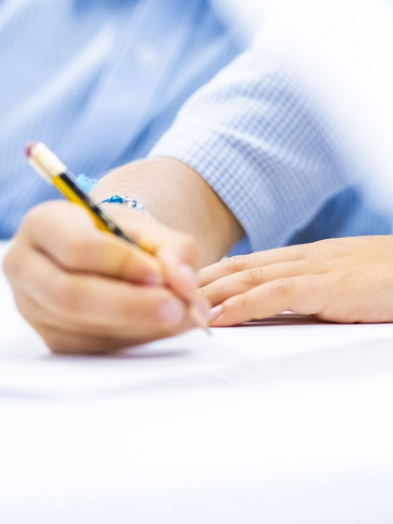 Child's hand writes with a pencil on white paper, wearing a blue checkered shirt in the background