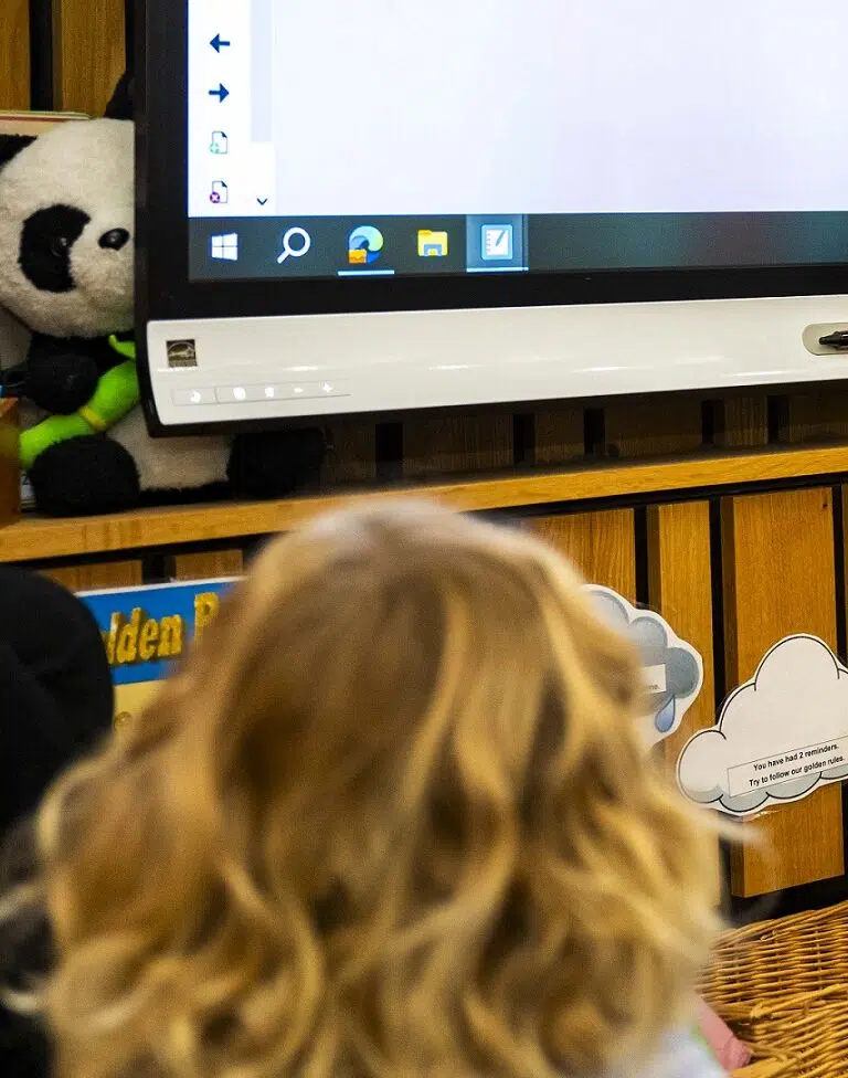 Child with blonde hair facing a computer screen in a classroom; panda plush on the shelf beside the monitor.