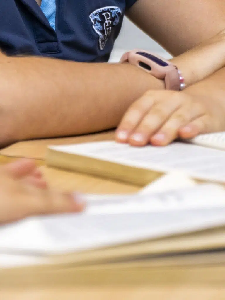 Close-up of a student with a blue polo shirt resting their hands on an open notebook at a desk.