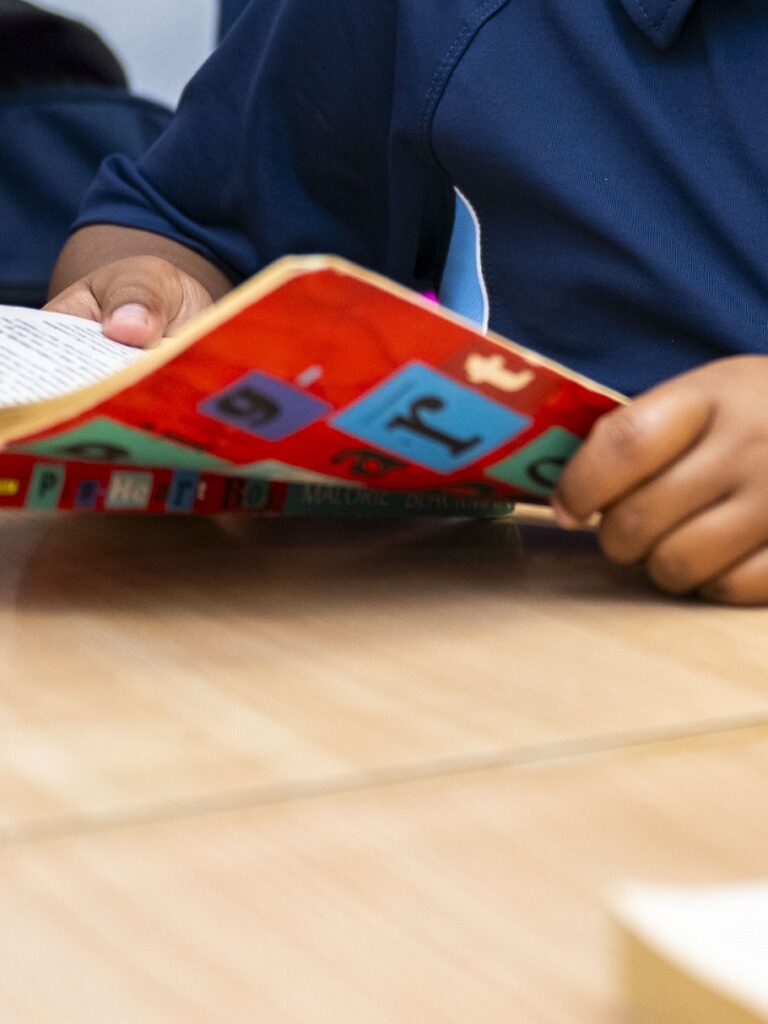 Child in a blue shirt reading a colorful sticker-covered book at a table, hands on the pages