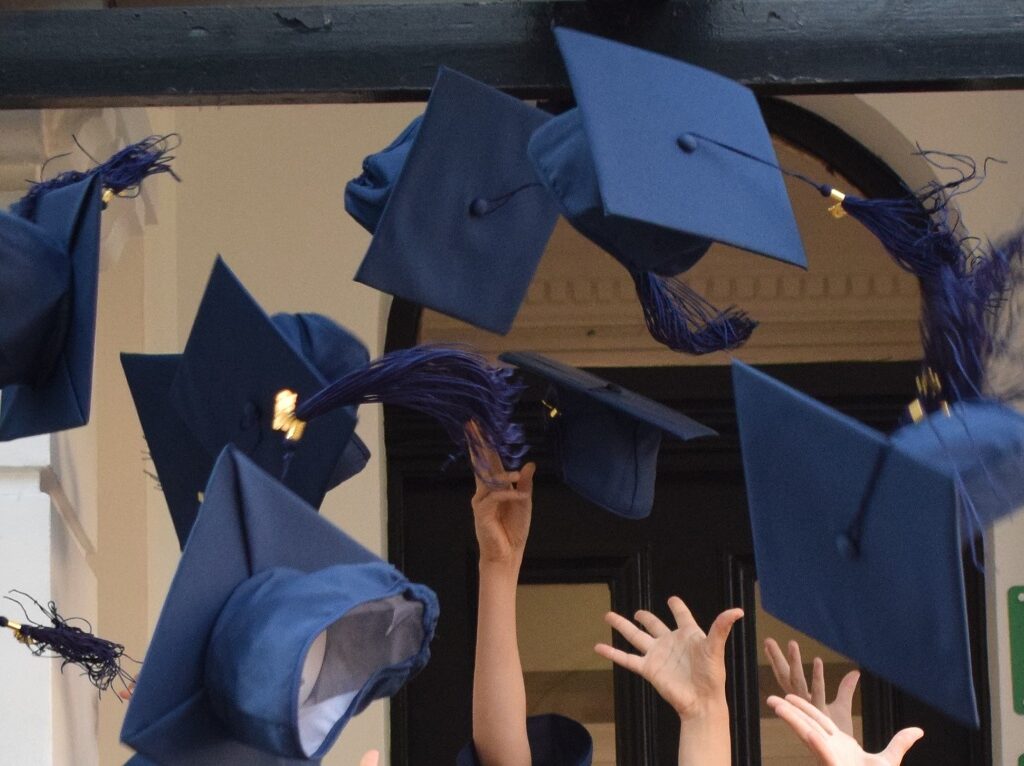 Blue graduation caps are being tossed in the air from a doorway, with hands reaching up in celebration.