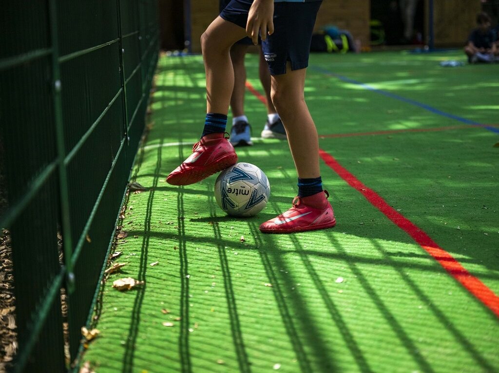 Child's red soccer shoes near a white ball on a green turf beside a fence during a game.