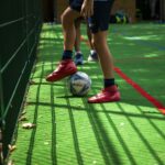 Child's red soccer shoes near a white ball on a green turf beside a fence during a game.