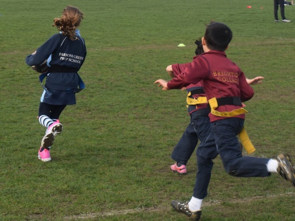 Two children in school uniforms run across a grassy field during a drill or race. The blue jacket reads 'Parsons Green Prep School' and the maroon jacket reads 'Brighton College'.