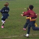 Two children in school uniforms run across a grassy field during a drill or race. The blue jacket reads 'Parsons Green Prep School' and the maroon jacket reads 'Brighton College'.