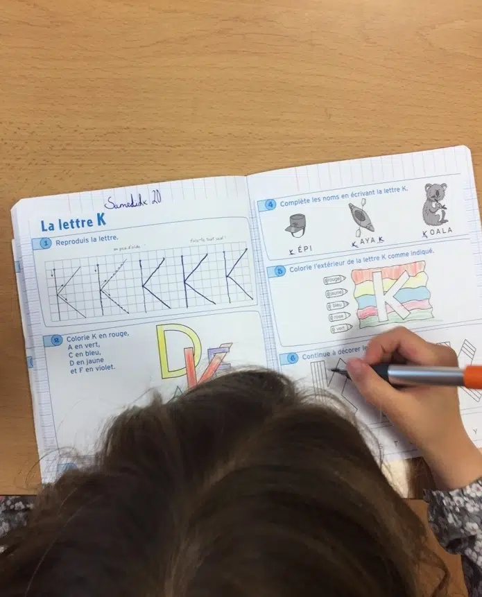 Top-down view of a workbook on a wooden desk showing tracing and coloring activities for the letter K; a child writes with an orange marker at the right page.