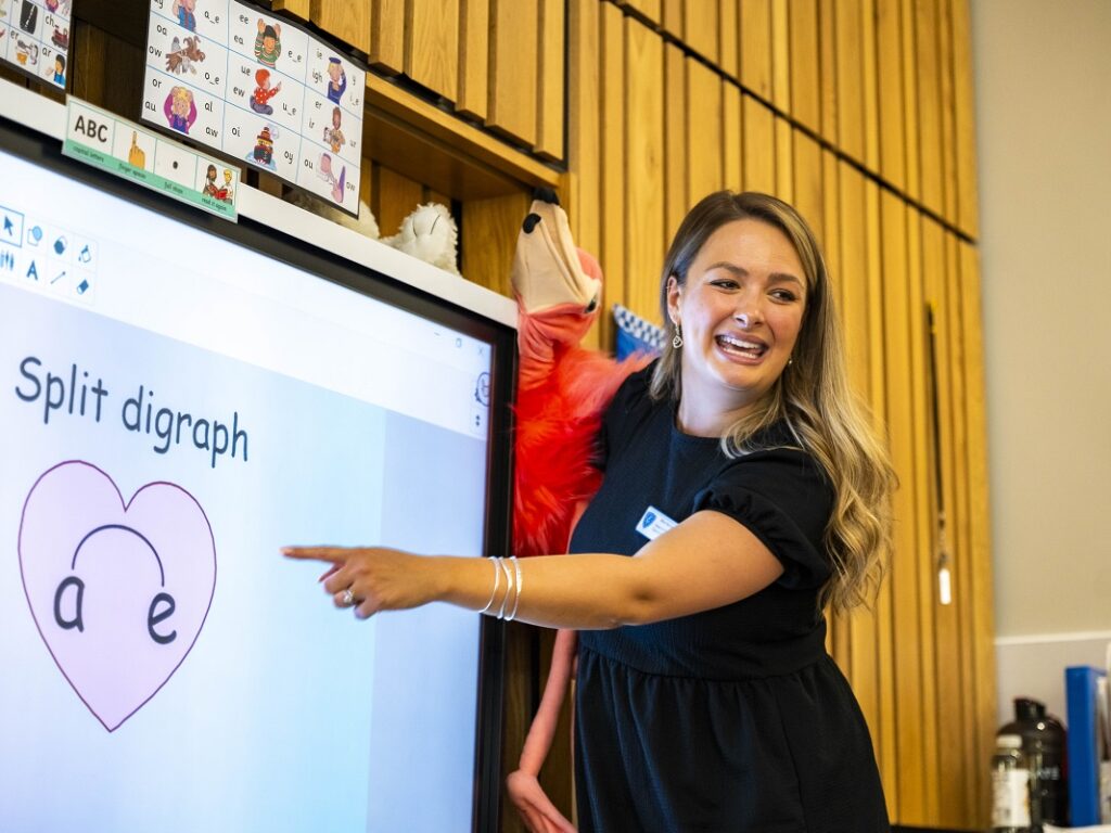 Teacher pointing at an interactive whiteboard that reads 'Split digraph' with a heart diagram labeled 'a' and 'e' in a classroom.