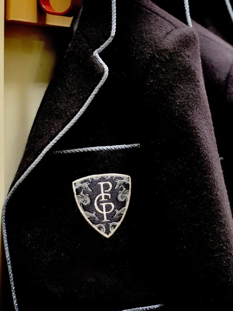 Close-up of a dark velvet blazer with pale blue piping and an embroidered shield crest on the pocket.