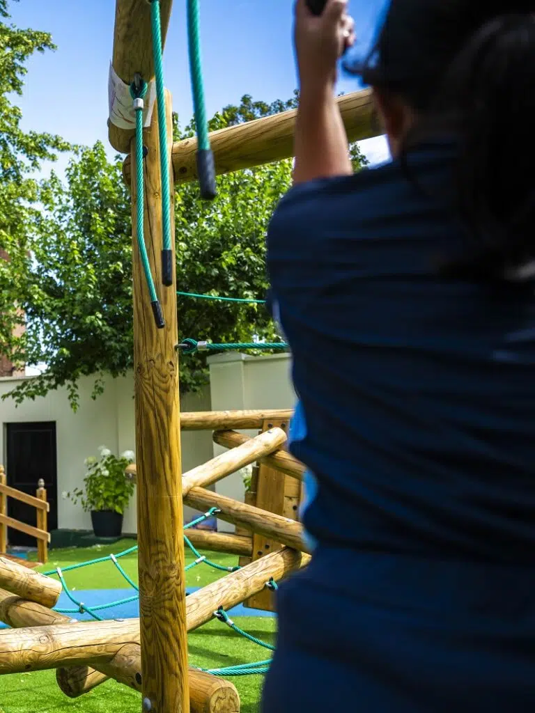 Back view of a person in a blue shirt climbing a wooden playground frame with green climbing ropes outdoors on a sunny day.
