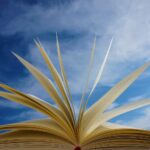 Open book with pages fanned upward, set against a bright blue sky, conveying imagination and learning