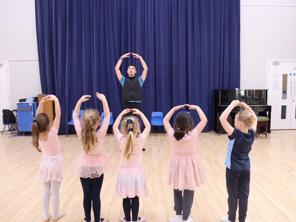 Dance instructor leading a ballet class as young girls raise arms in a circle pose in a studio with a blue curtain backdrop.