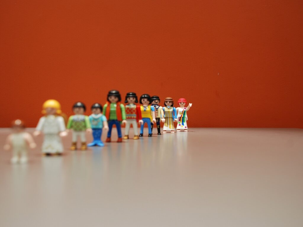 Line of colorful plastic figures (Playmobil) lined up on a table against an orange wall, left side slightly out of focus but clearly visible.
