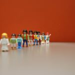 Line of colorful plastic figures (Playmobil) lined up on a table against an orange wall, left side slightly out of focus but clearly visible.