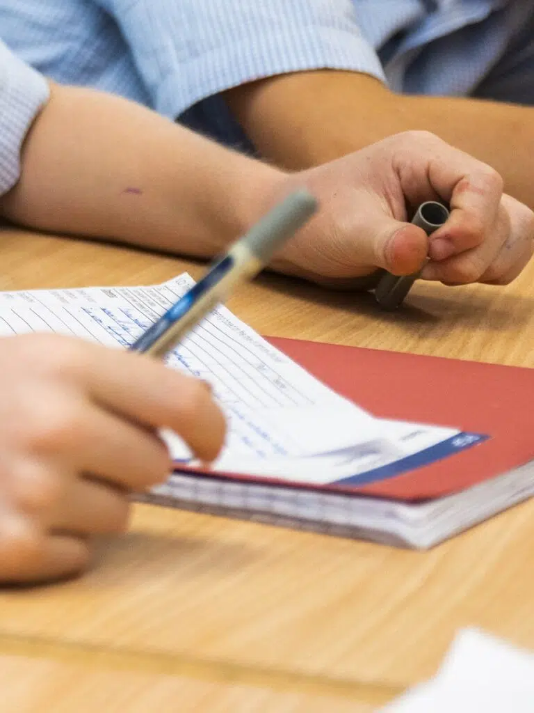 A student writes in a lined notebook with a blue pen while resting one arm on the desk, and a gray pen cap is held in the other hand.