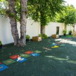 Playground area with numbered hopscotch squares 1–10 on a green surface, shaded by trees along a white wall.