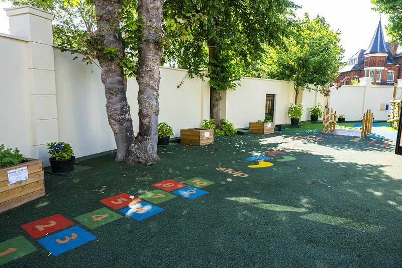 Playground area with numbered hopscotch squares 1–10 on a green surface, shaded by trees along a white wall.