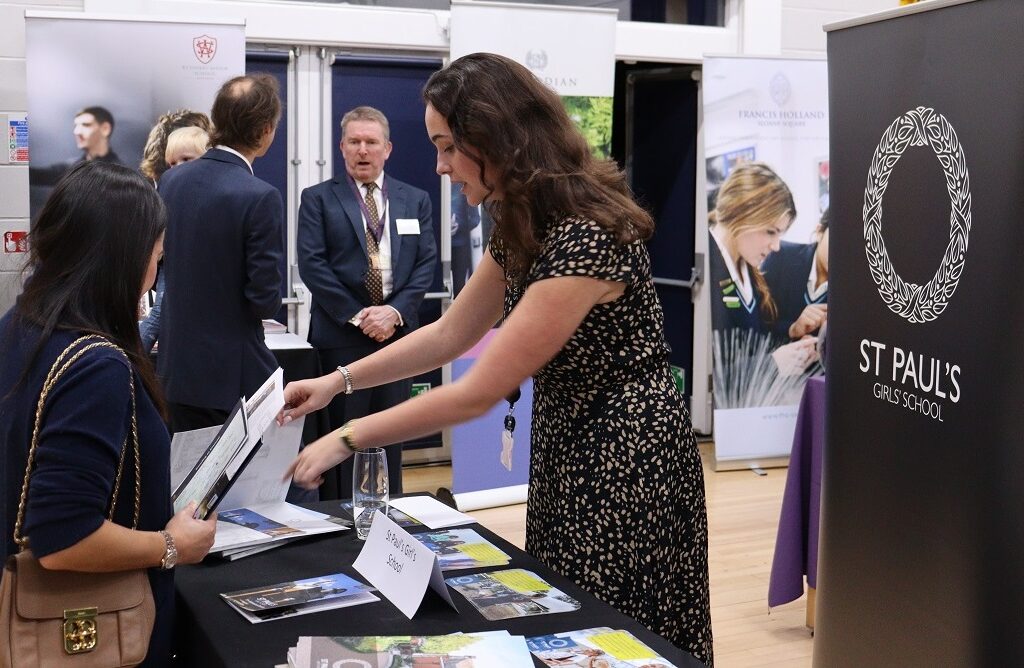 Woman in a black patterned dress hands brochures to a attendee at a school information fair, with a St Paul’s Girls’ School banner visible in the background.