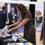 Woman in a black patterned dress hands brochures to a attendee at a school information fair, with a St Paul’s Girls’ School banner visible in the background.