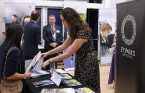 Woman in a black patterned dress hands brochures to a attendee at a school information fair, with a St Paul’s Girls’ School banner visible in the background.