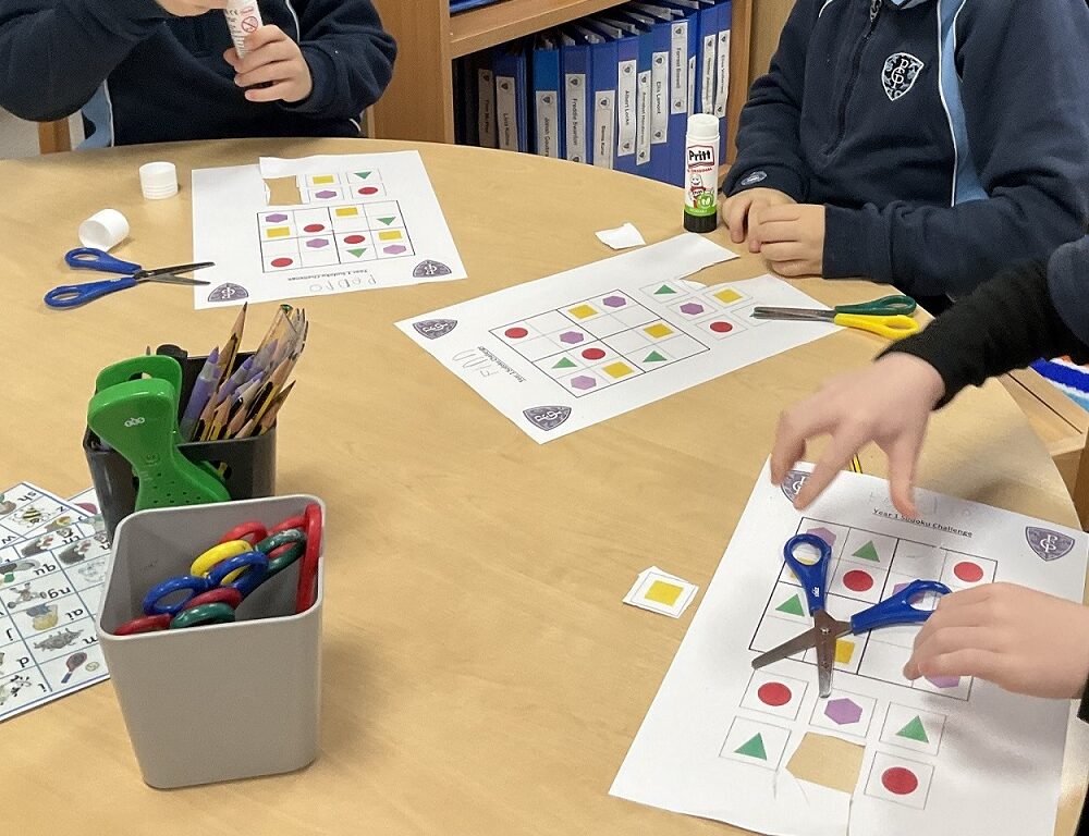 Children working on shape-matching worksheets at a table with scissors, glue, and colorful craft supplies nearby in a classroom setting.