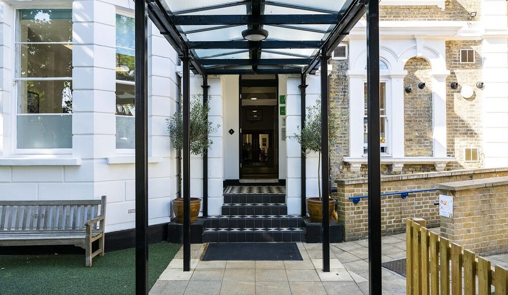 Entrance to a building with a glass canopy, black columns, and tiled steps flanked by potted plants.