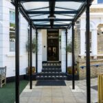 Entrance to a building with a glass canopy, black columns, and tiled steps flanked by potted plants.
