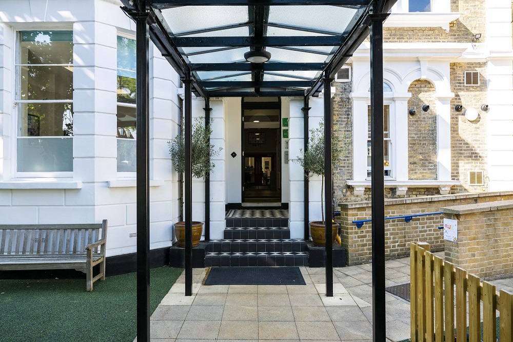 Entrance to a building with a glass canopy, black columns, and tiled steps flanked by potted plants.