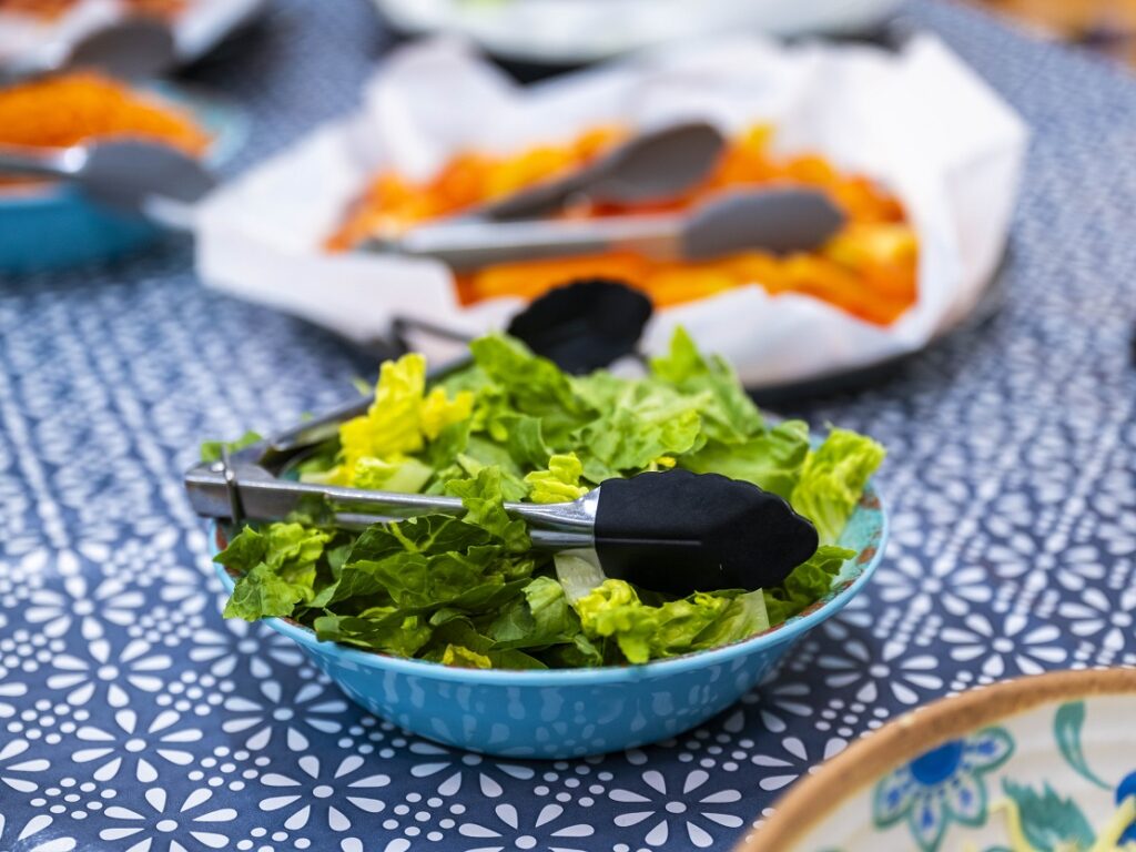 Blue bowl filled with fresh mixed greens and lettuce leaves, with metal tongs resting on top, on a blue patterned tablecloth at a buffet-style setup.