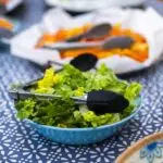 Blue bowl filled with fresh mixed greens and lettuce leaves, with metal tongs resting on top, on a blue patterned tablecloth at a buffet-style setup.