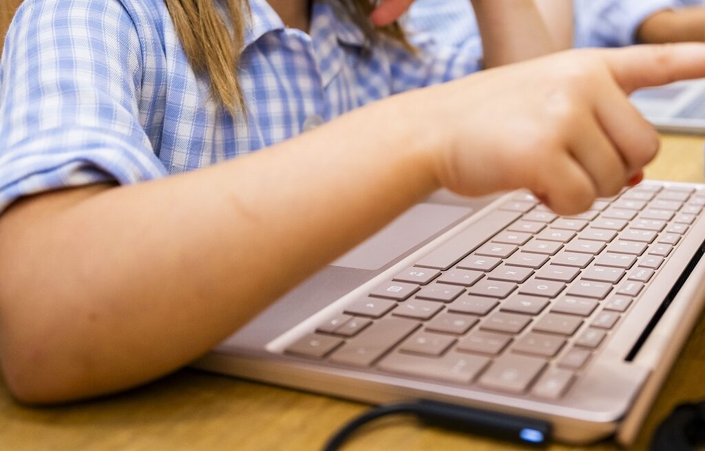 Child wearing a blue plaid shirt typing on a silver laptop at a wooden table in a classroom or study area on the keyboard area with hands in motion.