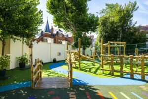 Colorful wooden playground structures with rope bridges and a blue path, shaded by trees in a sunny schoolyard or urban backyard setting.