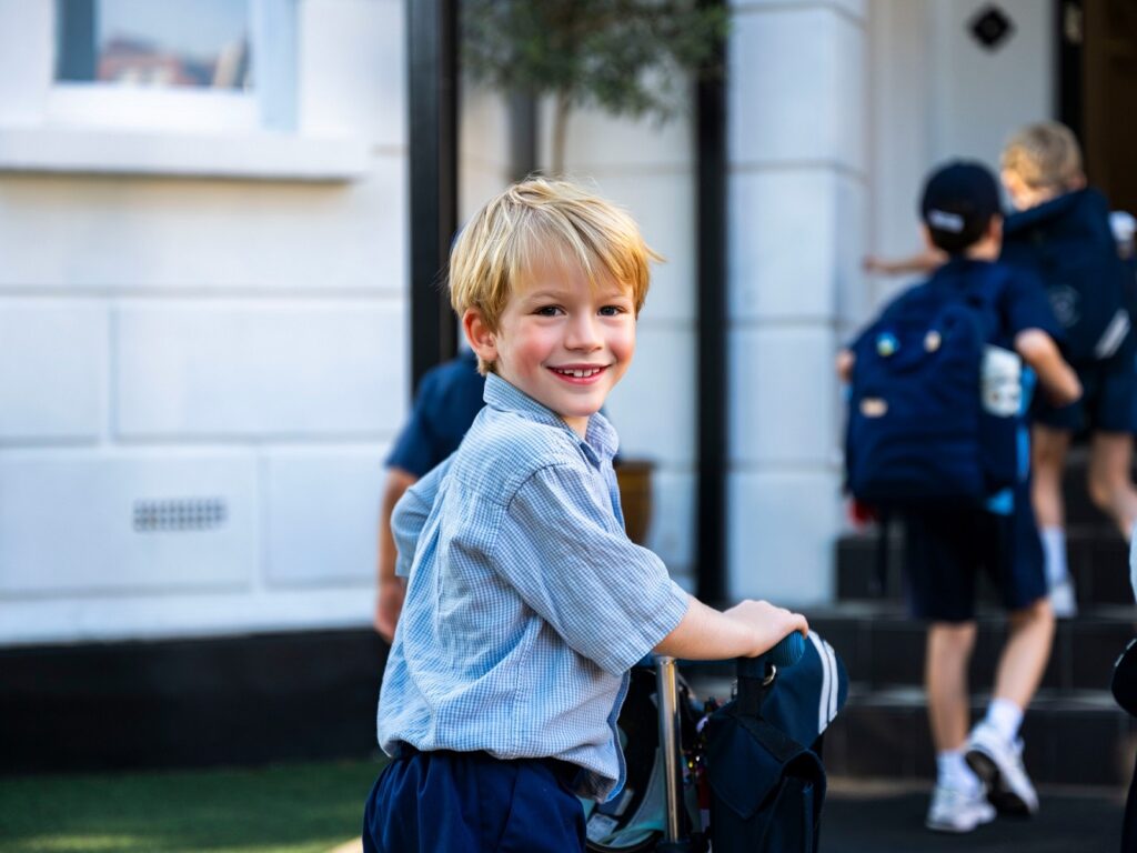 Smiling young boy in a light blue check shirt and navy shorts with a wheeled backpack, standing outside a white building.