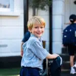 Smiling young boy in a light blue check shirt and navy shorts with a wheeled backpack, standing outside a white building.