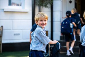 Smiling young boy in a light blue check shirt and navy shorts with a wheeled backpack, standing outside a white building.