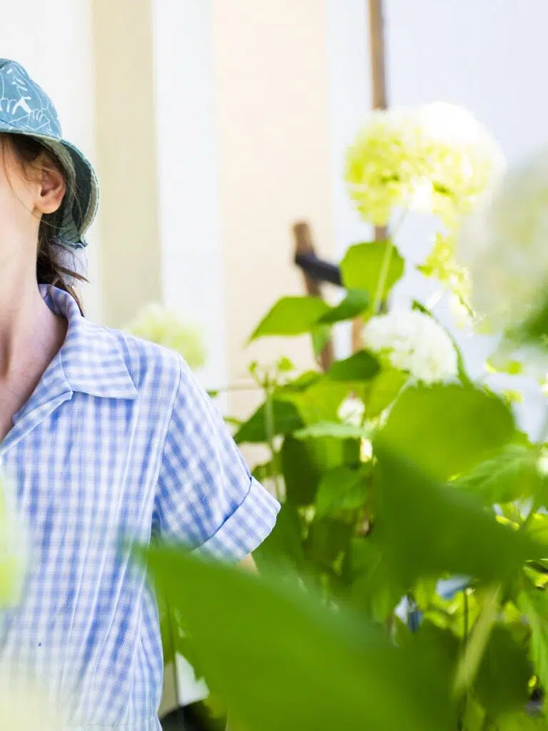Person wearing a blue checkered shirt and a blue hat standing among green plants and white flowers outdoors.