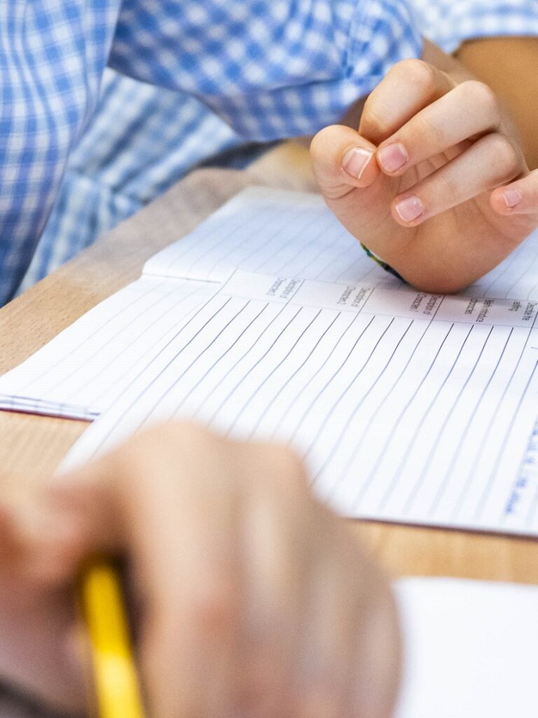 Close-up of a student's hand writing with a yellow pencil over a lined notebook on a wooden desk.