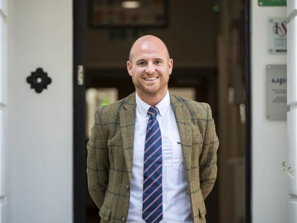 Smiling man in a brown checkered blazer, white shirt, and blue striped tie standing in a doorway.